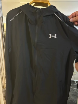 Svart vindjacka från Under Armour - Snygg svart vindjacka från Under Armour med huva och dragkedja framtill. Jackan har vita detaljer på axlarna och en vit logga på bröstet. Tillverkad i lätt och vindtätt material, perfekt för blåsiga dagar.