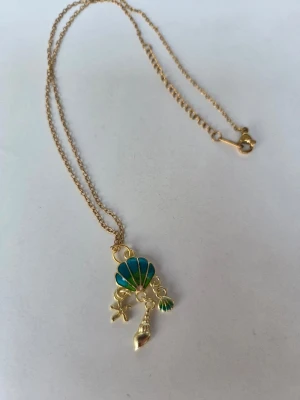 Guldigt halsband med snäckhänge - Snyggt halsband i guldfärgad metall med ett hänge som har en blågrön snäcka, en liten sjöstjärna och två små snäckor. Kedjan är tunn och har ett klassiskt lås, reglerbart upp till 45 cm. Halsbandet är monterat av mig.