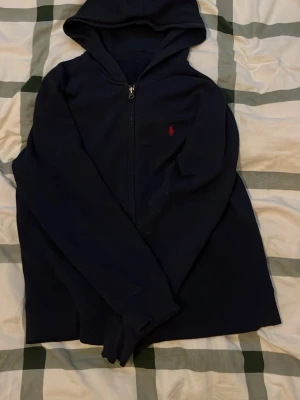 Mörkblå hoodie från Ralph Lauren - Mörkblå hoodie med dragkedja från Ralph Lauren. Klassisk design med huva och det ikoniska röda Polo-logotypen broderad på bröstet. Tillverkad i mjukt material som känns skönt mot huden. Perfekt för en avslappnad och stilren look.