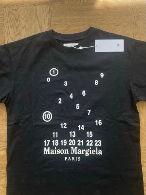 Maison Margiela T-shirt  - Cool svart t-shirt från Maison Margiela med ikoniskt tryck av siffror och logga framtill. Klassisk rak passform och rund halsringning. Tillverkad i mjuk bomull som känns skön mot huden. Perfekt för dig som gillar streetwear och designerplagg.