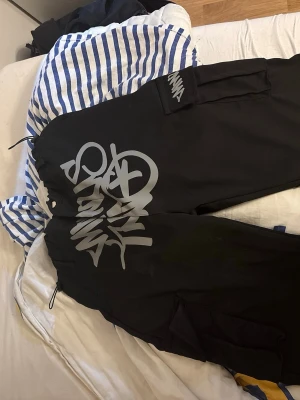Svarta mjukisbyxor Stay Wild - Svarta mjukisbyxor från Stay Wild med stor vit graffiti-inspirerad logga på benet. Byxorna har resår i midjan med dragsko och praktiska fickor på sidorna. Perfekta för en avslappnad och street-inspirerad stil.