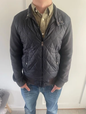 Svart quiltad cardigan - Säljer en svart quiltad med quiltat mönster på framsidan och släta ärmar. Jackan har dragkedja, tryckknappar vid kragen och två fickor med knappstängning. Materialet är troligtvis polyester och bomull, perfekt för en cool och avslappnad look. Jackan är lite solblekt 