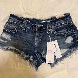 Mörkblå denimshorts Glamroot - Säljer ett par mörkblå denimshorts använda 1gång prislapparna är avtagna dem är från Ginatricot med slitna detaljer och fransig kant. Shortsen har låg midja, klassiska fem fickor och bälteshällor. Perfekta för varma dagar och en avslappnad look.