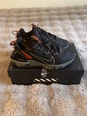 Nike React Vision svarta sneakers - Nike React Vision sneakers i svart med grå sula och orange detaljer. Skorna har en chunky design, mesh-överdel för bra andningsförmåga och reflekterande Swoosh på sidan. Färgglada snören och robust yttersula ger en sportig vibe.