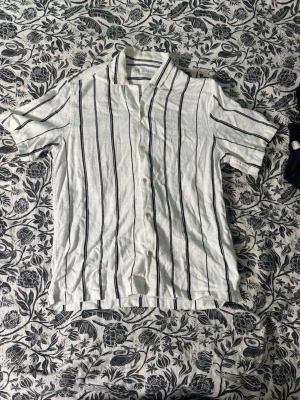 Vit randig linneskjorta från H&M - Säljer en vit skjorta i linne från H&M med svarta vertikala ränder. Skjortan har korta ärmar, klassisk krage och knäppning framtill. Perfekt för varma dagar och ger en avslappnad men stilren look.