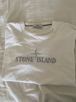 Vit Stone Island t-shirt med logga - Fläck går bort i tvätten innan frakt, lapparna borta pga stora och störiga! Snygg vit t-shirt från Stone Island med den klassiska loggan och kompassen tryckt i silvergrått på bröstet. T-shirten har rund hals och korta ärmar. Tillverkad i mjukt bomullsmaterial för en skön känsla.