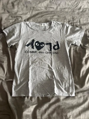 Grå Comme des Garçons PLAY t-shirt - Säljer en grå t-shirt från Comme des Garçons PLAY med det ikoniska hjärtlogotypen och tryck framtill. T-shirten har rund hals, korta ärmar och är gjord i mjuk bomull. Perfekt för dig som gillar streetwear och stilrena plagg.