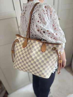 Louis Vuitton Damier Azur handväska - Snygg handväska från Louis Vuitton i klassiskt Damier Azur-mönster. Väskan har beige och vita rutor med detaljer i ljusbrunt läder och långa axelband. Perfekt storlek för att bära det viktigaste och ger en lyxig touch till din outfit.