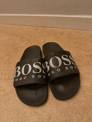 Svarta slides från Hugo Boss - Svarta sandaler från Hugo Boss med bred rem över foten och stor vit logga framtill. Tillverkade i syntetmaterial med räfflad sula för extra grepp. Perfekta för chill dagar eller att slänga på sig till och från gymmet.
