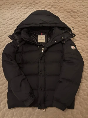 Svart dunjacka från Moncler - Snygg svart dunjacka från Moncler med stor huva och puffig design. Jackan har dragkedja och knappar framtill samt Moncler-logga på ärmen. Perfekt för kalla vinterdagar och riktigt varm tack vare dunfyllningen.
