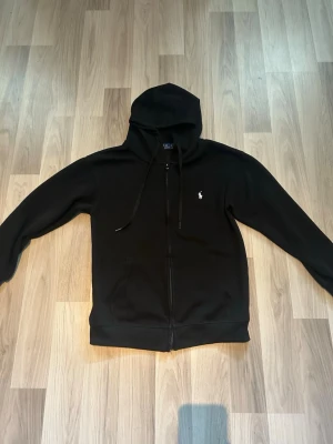 Svart zip hoodie från Polo Ralph Lauren  - Svart zip hoodie med dragkedja från Polo Ralph Lauren. Klassisk design med huva och snören, två fickor framtill och den vita loggan på bröstet. Jag säljer då den var lite liten för mig! Det är inge täcken på användning