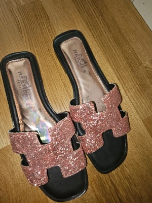 Glittriga sandaler rosa - Säljer ett par snygga sandaler med breda remmar täckta av roséguldglitter. Sandalerna har svart sula och öppen tå, samt en ikonisk H-formad design på ovandelen. Perfekta för dig som vill sticka ut med lite extra bling.