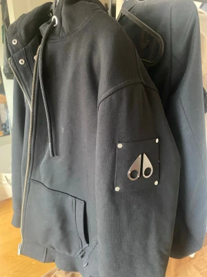 Svart hoodiejacka från Moose Knuckles - Svart hoodiejacka från Moose Knuckles med dragkedja, huva med snörning och stor huva. Inget kvitto till denna tyvärr men sitter äkthets tag på insidan utav jackan! Säljes då den inte används så speciellt mycket! Men i väldigt schysst skick☺️ bara att skriva vid nån fundering!