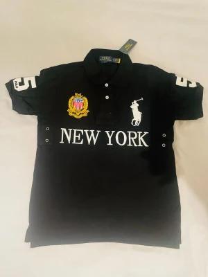 Svart pikétröja New York Polo Ralph Lauren - Svart pikétröja från Polo Ralph Lauren med vit logga och texten NEW YORK framtill. Broderat emblem i gult, blått och rött på bröstet samt siffran 5 på ärmarna. Klassisk krage och knappar framtill. Helt ny, storlek Small