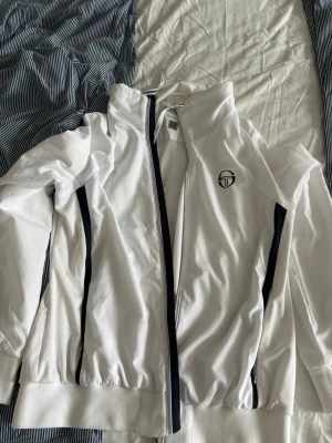 Vit träningsjacka från Sergio Tacchini - Säljer en vit träningsjacka från Sergio Tacchini med svarta detaljer längs dragkedjan och sidorna. Jackan har hög krage, hel dragkedja och är tillverkad i ett lätt, ventilerande material som passar perfekt till sport och träning.