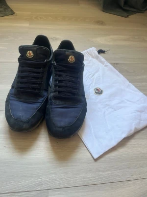 Mörkblå Moncler sneakers med mockadetaljer - Snygga svarta sneakers från Moncler med mörkblå textil och mockadetaljer. Klassisk låg modell med svart snörning och Moncler-logga på plösen. Skorna har en sportig siluett och kommer med en vit dustbag med logga.