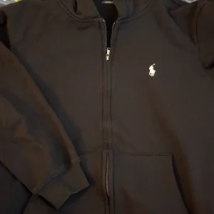 Svart hoodie från Polo Ralph Lauren - Svart hoodie från Polo Ralph Lauren i medium med klassisk vit logga broderad på bröstet. Tröjan har dragkedja framtill, huva och ribbade muddar vid ärmslut och nederkant. Custom fit-modell som är både stilren och bekväm.       Sitter som en Small