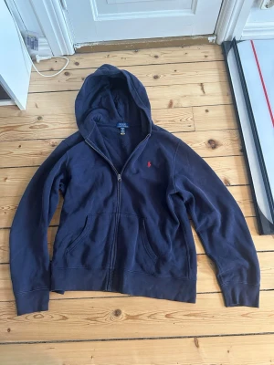Mörkblå hoodie från Polo Ralph Lauren - Mörkblå hoodie från Polo Ralph Lauren med dragkedja framtill och två stora fickor. Klassisk röd logga broderad på bröstet och mjukt material som känns skönt mot huden. Perfekt för en avslappnad och stilren look. Köpt på Nk i Stockholm för 1499 mitt pris 799. Storlek Xl i barnstorlek passar S jag är 180 och den är lite kort så mellan 170 och 178 är perfekt