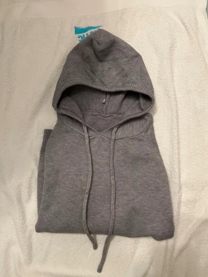 Grå merinoull hoddie - Mysig grå hoodie med huva och dragsko. Tillverkad i mjukt stickat material och har ribbade muddar vid ärmslut och nederkant. Perfekt för en avslappnad stil och enkel att matcha med jeans eller joggers. Den är Oanvänd!