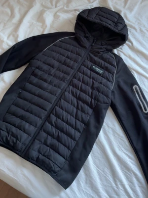 Svart cardigan från McKenzie - Snygg svart cardigan från McKenzie med vadderad framsida och huva. Jackan har dragkedja framtill, en ficka med dragkedja på ärmen och sportig design. Perfekt för dig som gillar streetwear och vill ha en lätt jacka till vardags.