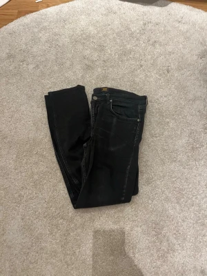 Svarta slim jeans från Lee - Säljer ett par svarta slim fit jeans från Lee med klassisk femficksdesign och knappgylf. Jeansen har en mörk tvätt och raka ben, perfekta för en clean och stilren look. Passar dig som gillar tidlös stil och enkelhet.