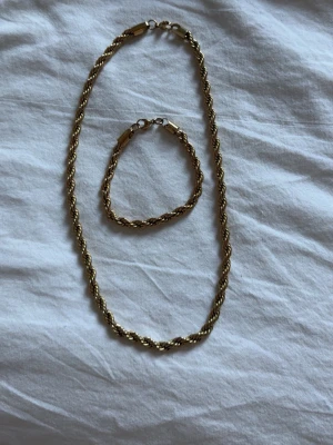 Guldfärgat halsband och armband set - Säljer ett matchande set med halsband och armband i guldfärgad metall. Båda har tvinnad kedja och klassisk karbinhake. Perfekt för dig som gillar stilrena accessoarer med lite extra bling.