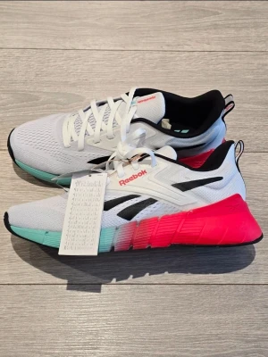 Reebok vita sneakers med färgade sulor - Snygga vita Reebok sneakers med mesh-ovandel, svarta detaljer och logga på sidan. Sulan är i en cool färgkombination av turkos, grått och starkt rosa. Skorna har vita snören och svart insida. Perfekta för dig som vill sticka ut med dina skor.