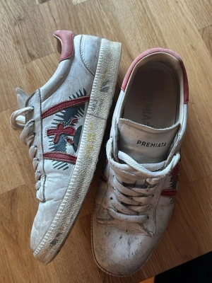 Premiata vita sneakers med röd detalj - Snygga vita sneakers från Premiata med röda och svarta detaljer på sidan samt röd hälkappa. Skorna har klassisk låg modell, snörning och coolt broderat mönster på sidan. Perfekta för dig som gillar streetstyle och vill sticka ut lite extra.