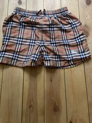 Rutiga shorts från Burberry - Säljer ett par klassiska rutiga shorts från Burberry i beige, svart, vitt och rött. Shortsen har elastisk midja med dragsko och sidofickor. Ikoniskt Burberry-mönster och logga på benet. Perfekta för en trendig och avslappnad stil. Jag säljer dom eftersom dom är för små för mig! Storleken säger medium men skulle säga att dom passar xs möjligtvis S. Jag köpte dom här på Plick för 800kr förra året, han jag köpte dom av sa att dom var äkta, men kan ej garantera de!