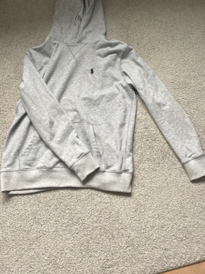 Grå hoodie från Polo Ralph Lauren - Snygg ljusgrå hoodie från Polo Ralph Lauren med klassisk huva och känguruficka. Tröjan har det ikoniska lilla mörkblå logobroderiet på bröstet och ribbade muddar vid ärmslut och nederkant. Perfekt för en chill och stilren look.
