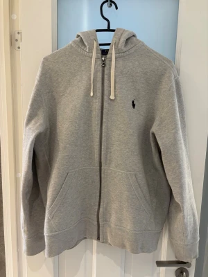 Grå zip hoodie från Polo Ralph Lauren - Klassisk grå hoodie med dragkedja från Polo Ralph Lauren. Har två fickor framtill och den ikoniska lilla loggan broderad på bröstet. Tillverkad i mjuk bomullsblandning som är skön att ha på sig. Perfekt för en chill och avslappnad stil.