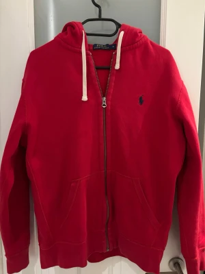 Röd hoodie från Polo Ralph Lauren - Riktigt snygg röd hoodie från Polo Ralph Lauren med dragkedja, huva med vita snören och kängurufickor. Klassisk blå logga broderad på bröstet. Tillverkad i mjuk bomull som känns skön mot huden. Perfekt för en avslappnad och stilren look. Dragkedjan åker ner lite av sig själv