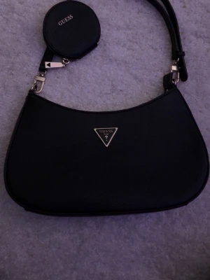 Svart axelremsväska från Guess - Snygg svart axelremsväska från Guess i konstläder med guldfärgade detaljer. Väskan har en klassisk halvmåneform, Guess-logga framtill och en liten rund myntväska som hänger på remmen. Baksidan har en praktisk dragkedjeficka. Den är i nytt skick och har bara använts en gång (Självklart äkta!) Skriv vid intresse skickar samma dag eller dagen efter!