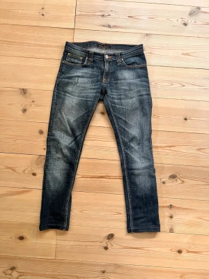 Blå slim fit jeans från Nudie Jeans - Säljer ett par blå slim fit jeans från Nudie Jeans med snyggt slitna detaljer och klassisk femficksmodell. Jeansen har en något tvättad look och orange sömmar. Wide 28 Lenght 30