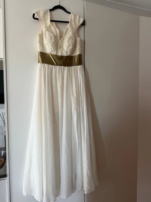Vit lång balklänning med pärldetaljer - Elegant vit långklänning med v-ringad överdel dekorerad med pärlor och broderade blommor. Midjan markeras av ett brett guldigt band och kjolen faller mjukt i ett tunt, svepande tyg. Perfekt för speciella tillfällen där du vill sticka ut. Obs se mitten bild för en liten fläck, man tänker inte på den så mycket. 