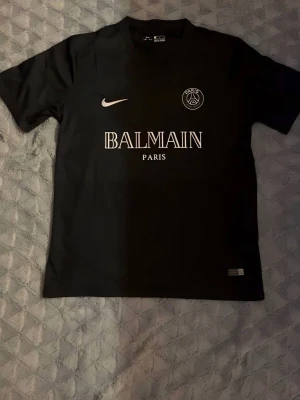 Svart PSG x Balmain tröja Nike - Snygg svart PSG x Balmain tröja från Nike med vita tryck på bröstet. Klassisk rund hals, korta ärmar och Dri-FIT material som andas. Perfekt för fotboll eller streetwear. Stora Balmain-loggan och PSG-emblemet framtill.