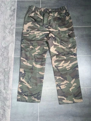 Klassiska camo cargobyxor - Säljer ett par cargobyxor i klassiskt militärgrönt camomönster. Byxorna har flera stora fickor på sidorna och baktill, samt elastisk midja för skön passform. Perfekta för dig som gillar streetwear och utility-stil.