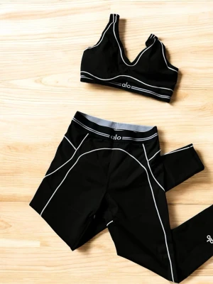Svart träningsset från Alo Yoga - Snyggt svart träningsset från Alo Yoga med sport-bh och tights. Setet har vita kontrastsömmar och logga på både bh och byxa. Tillverkat i stretchigt funktionsmaterial som passar perfekt till gymmet eller yoga.