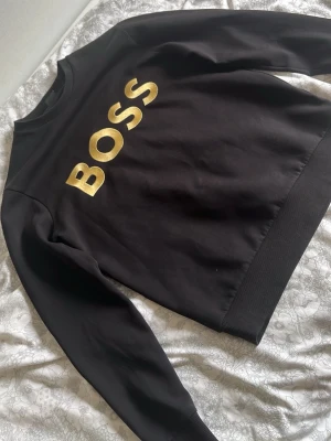 Svart sweatshirt från BOSS  - Säljer en svart sweatshirt från BOSS med stor guldfärgad BOSS-logga på bröstet. Tröjan har rund halsringning, långa ärmar och ribbade muddar vid ärmslut och nederkant. Tillverkad i mjukt material som känns skönt mot huden. Perfekt för en clean och stilren look.