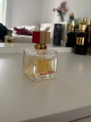 Valentino parfym i glasflaska - Säljer en lyxig parfym från Valentino i en fyrkantig glasflaska med gulddetaljer och röd accent på korken. Flaskan har en elegant design med Valentino-loggan i rött framtill. Perfekt för dig som gillar exklusiva dofter och snygg inredning på sminkbordet.