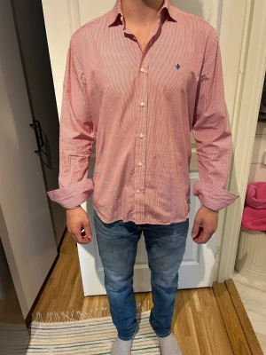 Rödvit randig skjorta från Morris - Klassisk rödvit randig skjorta från Morris med knappar framtill och liten broderad logga på bröstet. Skjortan har lång ärm, normal passform och är tillverkad i bomull. Perfekt för en stilren och avslappnad look.