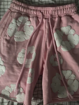 Rosa shorts med vita blommor - Säljer ett par rosa shorts med vita blommönster och resår i midjan samt snörning. Shortsen har fickor på sidorna och en bakficka. Perfekta för sommarens chill dagar och har en loose passform.