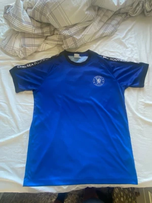 Blå Chelsea FC t-shirt - Snygg blå t-shirt från Chelsea FC med klubbens logga på bröstet och texten 'CHELSEA FOOTBALL CLUB' på axlarna. T-shirten har svarta detaljer vid krage och ärmslut och är tillverkad i ett lätt och sportigt material, perfekt för träning eller vardag.   Träningströja köpt i Chelseas officiella matchbutik under oktober 2019!