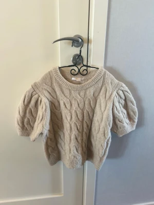 Beige stickad tröja Gina Tricot - Säljer en beige stickad tröja från Gina Tricot med korta puffärmar och kabelstickat mönster. Tröjan har rund halsringning och ribbade kanter. Perfekt för dig som gillar en mysig och trendig look.