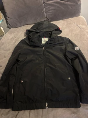 Moncler windbreaker  - Snygg svart vindjacka från Moncler med huva och dragkedja framtill. Jackan har två fickor med tryckknappar och Moncler-logga på vänster ärm. Tillverkad i ett lätt och vattenavvisande material, perfekt för blåsiga dagar.