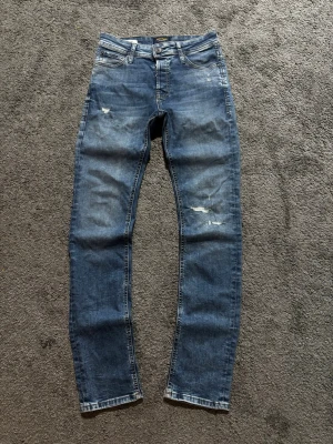 Jack & Jones jeans  - Snygga blå slim fit jeans med slitningar och distressed detaljer på benen. Klassisk femficksmodell med dragkedjegylf och knapp. Perfekta för en avslappnad och trendig look.