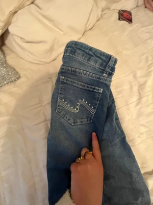 Blå bootcut jeans med nitar - Säljer ett par blå bootcut jeans med snygga nitar på bakfickan och klassisk femficksdesign. Jeansen har en ljus tvätt och raka ben, perfekta för en avslappnad men trendig look. Materialet är klassisk denim i bomull.