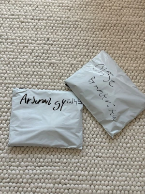 Fotbollströjor - Säljer två gråa plastpåsar med handskriven text på. Perfekta för förvaring eller frakt av mindre saker. Påsarna är rektangulära och gjorda av tunn plast.