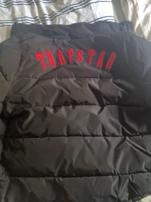 Svart dunjacka från Trapstar - Säljer en svart dunjacka från Trapstar med stor röd logga på ryggen och ett runt märke på bröstet. Jackan har en puffig design och är perfekt för kalla vinterdagar. Materialet är slitstarkt och håller värmen bra. Snygg streetwear-stil!