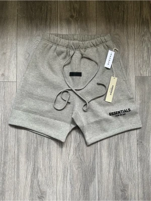 Grå Essentials shorts FÖRBESTÄLLING ENDAST  - Säljer ett par grå shorts från Essentials med snörning i midjan och tryckt logga på benet. Shortsen har en avslappnad passform och är perfekta för chill dagar. Tillverkade i mjuk bomull för maximal komfort. Helt nya,Fraktar inom 24 timmar 